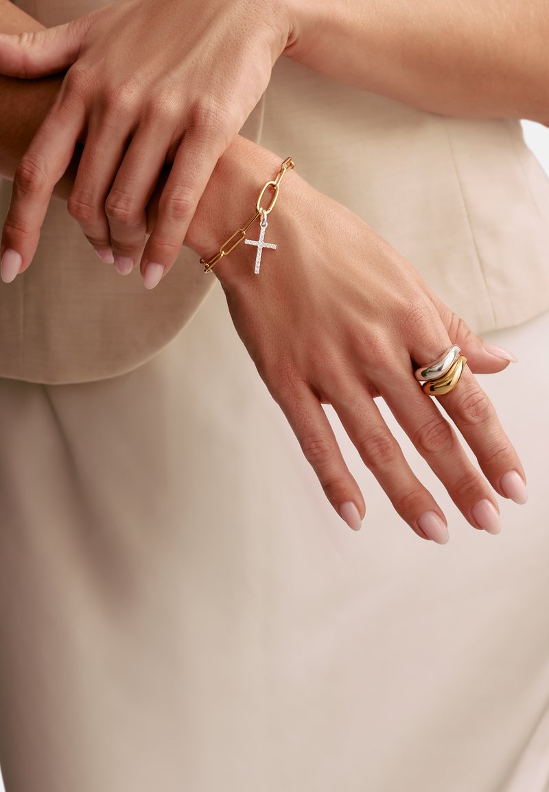 Bracelet chaîne en or avec un pendentif croix en argent sur un poignet ; plusieurs bagues en argent, en or et avec un motif torsadé aux doigts.