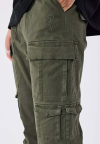 Close-up van olijfgroene cargobroek met klepzakken en geborduurde initialen op het dijbeen, hand rustend in de zak.