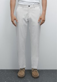 Lichtgrijze broek van gladde stof, met een knoopsluiting, steekzakken en een tapered fit. Gecombineerd met bruine suède schoenen.