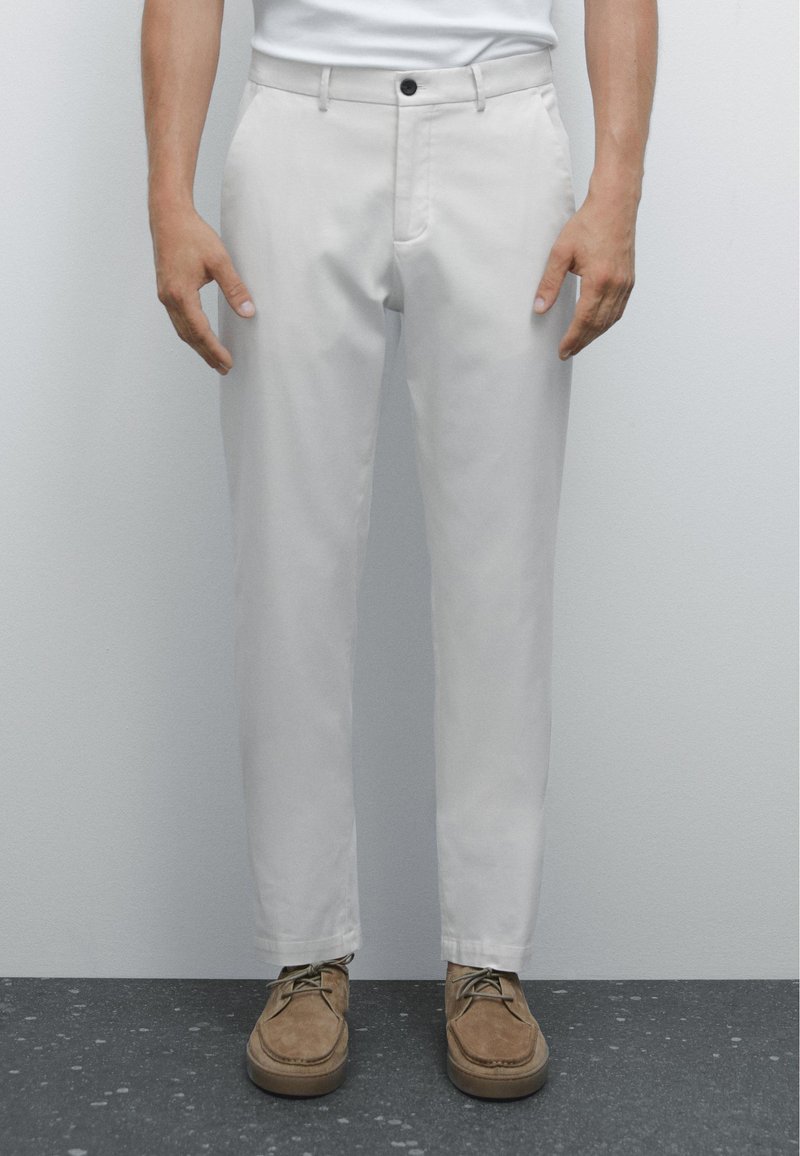 Lichtgrijze broek van gladde stof, met een knoopsluiting, steekzakken en een tapered fit. Gecombineerd met bruine suède schoenen.