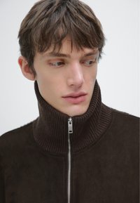 Brązowa zamszowa kurtka z prążkowanym wysokim kołnierzem, z metalowym zamkiem błyskawicznym z przodu i teksturowanymi detalami na kołnierzu.