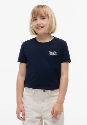 Barn med blond bob-frisure iført en mørkeblå Tommy Hilfiger t-shirt og lyse beige bukser, smilende mod en hvid baggrund.