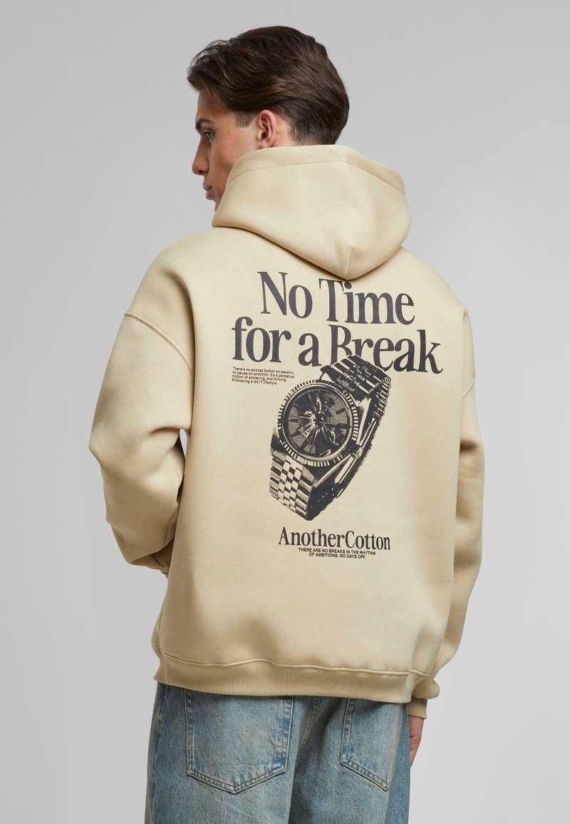 Beige hoodie met een grote zwarte grafiek op de achterkant die een horloge en tekst afgebeeld. Heeft een capuchon met trekkoord en ribgebreide zoom en manchetten.
