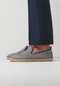 Espadrilles à enfiler à carreaux bleu marine et blancs avec une semelle en jute, présentant des découpes angulaires et un contour bleu le long des bords.