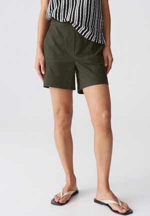 Personne portant un short olive arrivant aux genoux, un débardeur rayé noir et blanc, et des tongs beiges, debout sur un fond uni.