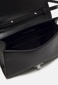 Čierna kožená kabelka s hladkou textúrou, elegantným dizajnom, vnútornými vreckami a kovovým zapínaním s brandingom Calvin Klein.