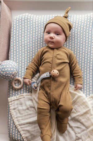 En baby i en stripete mustard-farget romper med en matchende knyttet lue. Omgitt av et mønstrete teppe, treleker og en smokk.