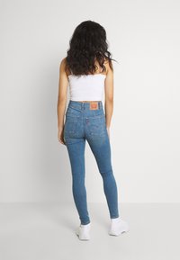 Vaqueros azules de talle alto con corte ajustado, con bolsillos traseros y un parche de cuero con la marca, combinados con una camiseta sin mangas blanca y zapatillas blancas.