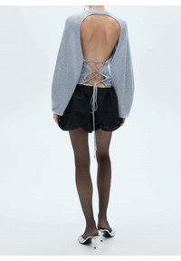 Frau trägt einen grauen Pullover mit offenem Rücken und Schnürdetail, schwarze Shorts, durchscheinende schwarze Strumpfhosen und weiße High Heels, steht vor einem weißen Hintergrund.