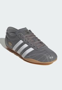 Γκρι δερμάτινο αθλητικό παπούτσι με τρεις λευκές ρίγες, σόλα από καουτσούκ χρώματος καραμέλας, ανάγλυφο λογότυπο στη γλώσσα και λεπτομέρειες με ραφές καπιτονέ.