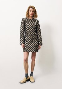 Hemdjurk met korte mouwen in beige met zwart bloemenpatroon, voorzien van een rechte snit en ronde halslijn. Gecombineerd met beige instappers en grijze sokken.