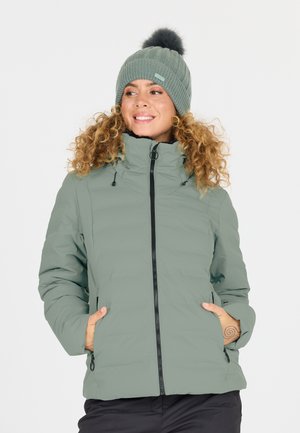 Kvinde med krøllet hår iført en lysegrøn dunjakke og matchende hue med pompom, smilende og kigger til siden.