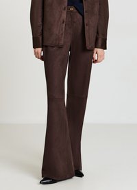 Braune Schlaghosen aus weichem, wildlederähnlichem Material mit glatter Textur. Kombiniert mit einer passenden, geknöpften Jacke und spitzen schwarzen Schuhen.