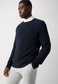 Marineblå ribbestrikket genser med lange ermer og rund hals, kombinert med lysegrå bukser. Stoffet ser ut til å være teksturert og varmt.