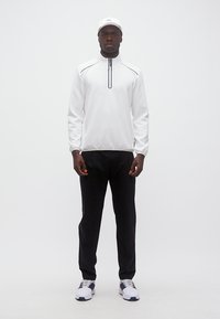 Άνδρας που στέκεται φορώντας λευκό πουλόβερ με μισό φερμουάρ, μαύρο παντελόνι, λευκό καπέλο και λευκά παπούτσια με μαύρες λεπτομέρειες πάνω σε απλό λευκό φόντο.