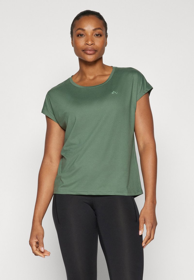 Woman wearing a short-sleeve green athletic shirt and black leggings, standing against a plain white background.