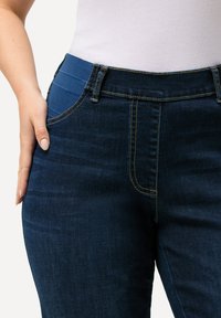 Tummansiniset farkut, joissa on ribbipintainen sininen vyötärö, etutaskut ja kontrastivärinen tikkaukset saumoissa ja taskuissa.