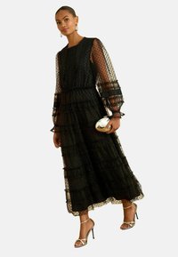 Čierna maxi šaty s poschodeným strihom a bodkovaným vzorom, priesvitnými dlhými rukávmi, riasenými vrstvami, elastickým pásom, skombinované s metalickou kabelkou a lodičkami.