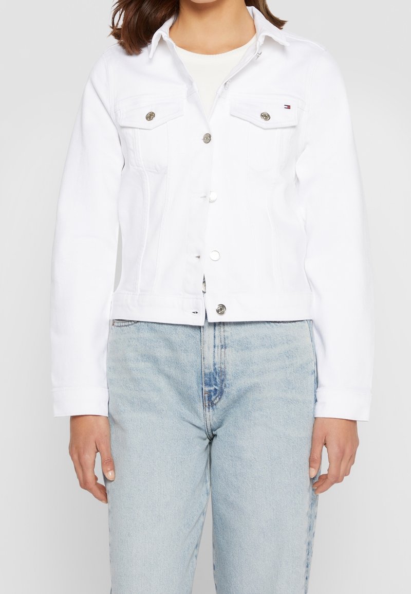 Femme portant une veste en jean blanche boutonnée sur un haut blanc avec un jean taille haute bleu clair, debout devant un fond uni.