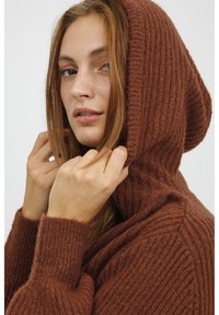 Felpa marrone lavorata a maglia con una texture a coste, maniche lunghe e un grande cappuccio. Il materiale sembra morbido e accogliente.