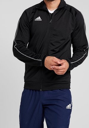 Mężczyzna noszący czarną kurtkę Adidas zapinaną na suwak z białymi paskami na rękawach oraz granatowe spodnie Adidas z logo na udzie.