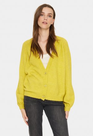 Geel vest met een gestructureerde brei, voorzien van een V-hals, vijf knopen en losvallende mouwen. Gecombineerd met donkere jeans.