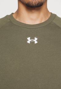 Gros plan d'une personne portant un sweat-shirt vert olive à col rond avec un logo Under Armour blanc sur la poitrine.