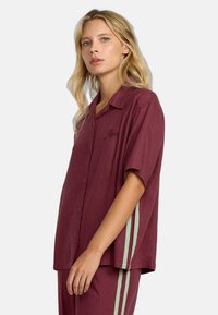 Camicia corta a maniche corte color marrone scuro con vestibilità rilassata, chiusura con bottoni sul davanti, dettagli ricamati e strisce verticali verdi sui lati.