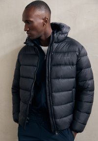 Giacca a vento nera con chiusura a zip e cappuccio. Presenta sezioni trapuntate, una superficie liscia e un logo discreto sul petto.