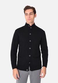 Cardigan lavorato a maglia nero con colletto alto, chiusura frontale a bottoni e due tasche anteriori. La texture è spessa, con polsini e orlo a coste.