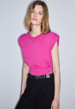 Junge Frau mit schulterlangem blondem Haar, die ein ärmelloses, leuchtend pinkes Top und schwarze Hosen mit einem schwarzen Gürtel trägt, stehend vor einer schlichten Wand.