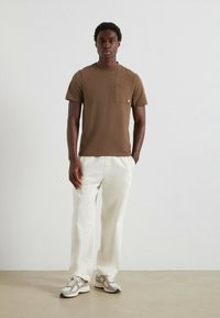 Maglietta corta marrone a maniche corte con tasca sul petto, abbinata a pantaloni bianchi a gamba larga. L'outfit è completato da scarpe da ginnastica grigie e beige.