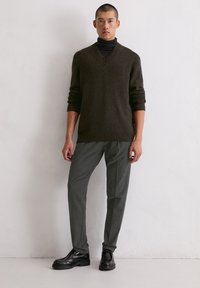 Dunkelgrüner Strickpullover mit V-Ausschnitt, kombiniert mit grauen, maßgeschneiderten Hosen und schwarzen Lederschuhen. Schlichtes Design, taillierte Silhouette.