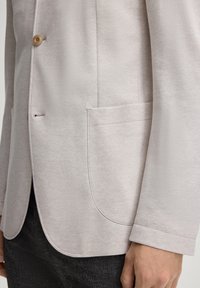 Blazer beige chiaro realizzato in tessuto testurizzato, con chiusura a un bottone e tasca laterale, esposto su uno sfondo neutro.