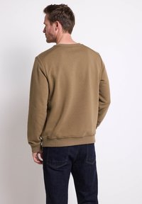 Felpa verde oliva con scollatura a girocollo, polsini e orlo a coste. Indossata con jeans blu scuro, che mostrano un tessuto semplice e strutturato.