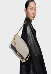Mujer con abrigo de cuero negro llevando un bolso TOUS beige y negro con correa de cadena dorada al hombro frente a un fondo blanco.