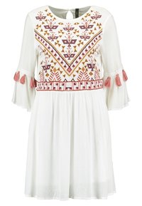 Vestido blanco con una tela texturizada, que presenta bordados geométricos coloridos y borlas rosas en las mangas acampanadas. Detalle de abertura en la espalda.