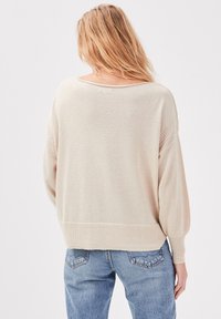 Nainen, jolla on pitkät vaaleat hiukset, pukeutuneena löysään vaaleanbeigeen neulepaitaan ja sinisiin farkkuihin, näkyy selkäpuolelta tasaisen vaaleaa taustaa vasten.