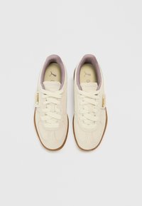 Zapatillas deportivas de ante beige con acentos en violeta claro, cordones planos y una suela de goma texturizada color caramelo. Presentan una pequeña etiqueta de la marca en el lateral.