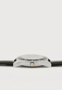 Στρογγυλό ρολόι χειρός με μαύρο καντράν, ασημένιο στερέωμα από ανοξείδωτο ατσάλι και μαύρο δερμάτινο λουράκι. Διακριτά σημάδια και κορώνα.