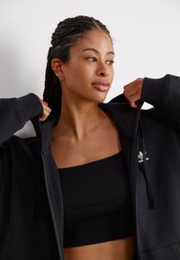 Juodas užtrauktukas švarkas, pagamintas iš minkštos medžiagos, su guma apiniais rankovėmis, dėvimas ant juodo raukšlėto maikės tipo marškinuko, su mažuoju baltu logotipu ant krūtinės.