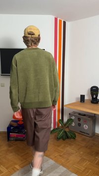 Olivově zelený pletený svetr, volný střih; hnědé šortky; bílé ponožky s teniskami. Jasně pruhovaná zeď; dřevěná podlaha; dekorativní rostlina.