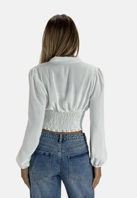 Weißes, kurzes Blusenshirt mit langen Puffärmeln und einem gesmokten Rücken. Kombiniert mit dunkelblauen, taillierten Jeans. Glatte Stofftextur.