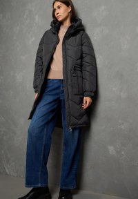 Donna che indossa un lungo cappotto imbottito nero, un maglione beige, jeans a gamba larga blu e stivali neri, in piedi contro un muro grigio texturizzato.
