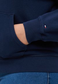 Sudadera azul marina con puños y dobladillo acanalados. Cuenta con un bolsillo frontal y un pequeño logo en rojo, blanco y azul en la manga.