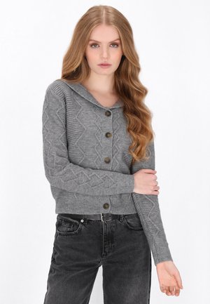 Ung kvinde med langt bølget hår iført en grå mønstret knapcardigan og sorte jeans mod en hvid baggrund.