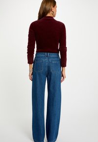 Donna in piedi di spalle, con indosso un maglione bordeaux a maniche lunghe con polsini arrotolati e pantaloni larghi a vita alta blu.