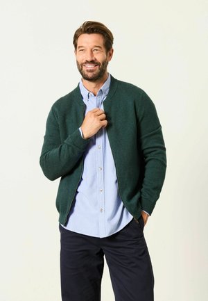Mann mit Bart, der einen grünen Reißverschluss-Pullover über einem hellblauen Hemd und dunklen Hosen trägt, lächelnd mit einer Hand auf dem Pullover.
