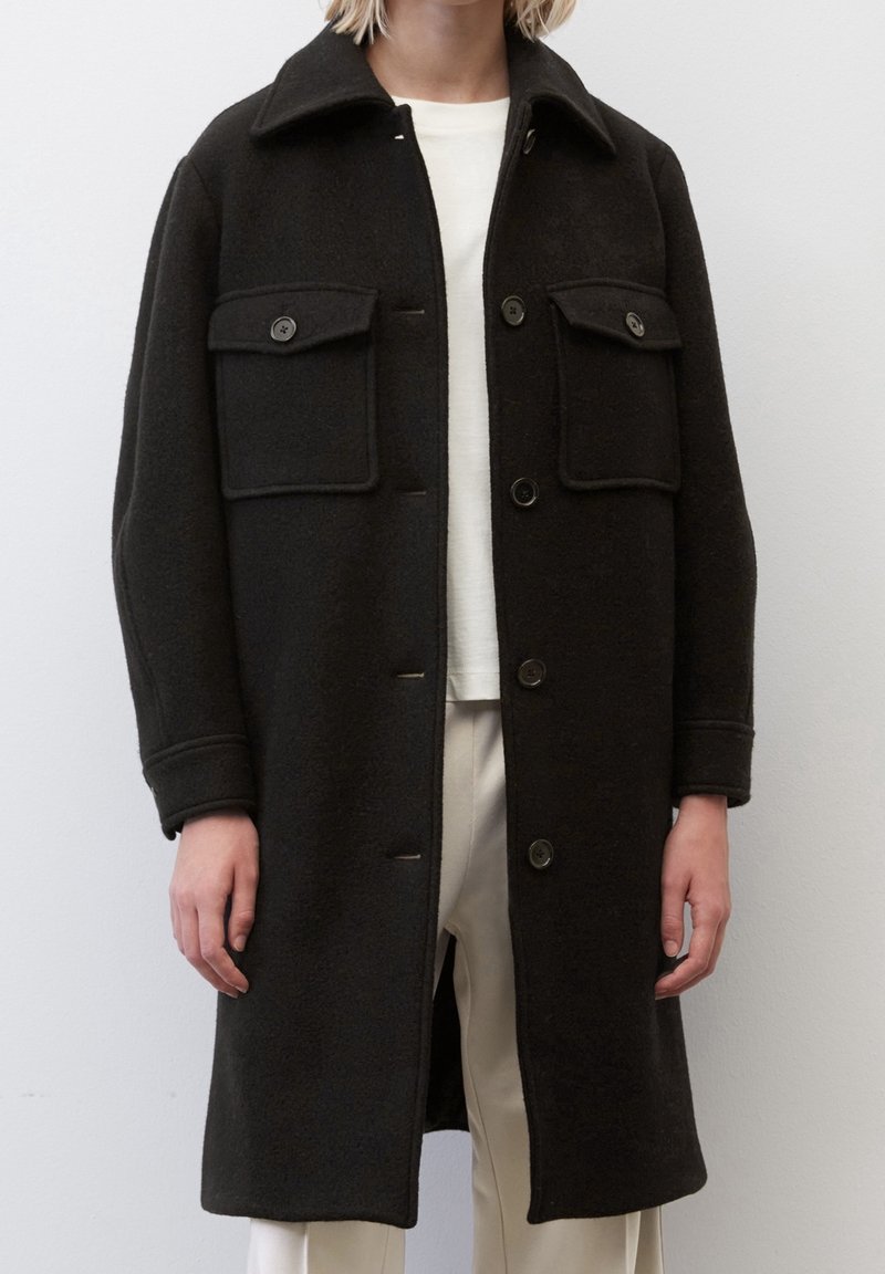 Cappotto in lana nera con vestibilità su misura, caratterizzato da un colletto a punte, due tasche frontali e chiusura a bottone. Tessuto liscio, design lungo fino al ginocchio.