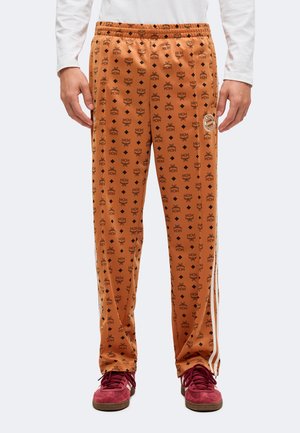 Modèle portant un pantalon de survêtement orange MCM à monogramme, avec des bandes latérales, et des baskets bordeaux sur un fond uni.